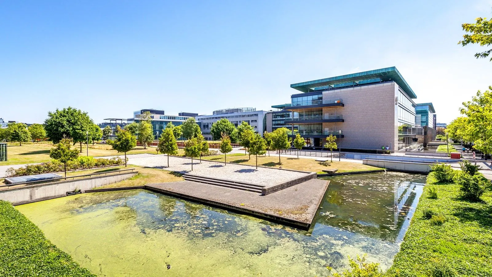 Modern kantoorgebouw aan het water in Kessler Park met groenstroken en bomen op een zonnige dag.